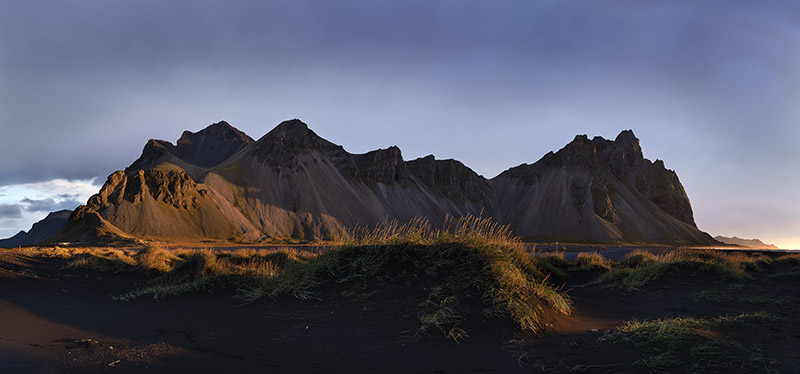 Fine art panoramic landscape photography of the gabbro rock peaks of Mount Vestrahorn at sunrise. The golden morning light illuminates the sweeping volcanic black sand dunes and green grasses of the Stokksnes peninsula in Iceland under a soft pastel sky.