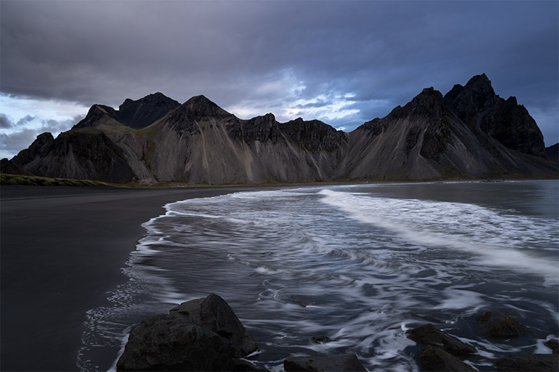 Fine art landscape photography of Mount Vestrahorn under a moody storm sky in Iceland. The dark, jagged peaks rise sharply over a black volcanic sand beach where breaking ocean waves create white surf and foam patterns in the foreground.