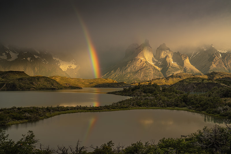 A dramatic golden hour view of Torres del Paine peaks with a vibrant rainbow reflecting in a calm lake.